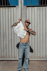 Chico joven musculado y tatuado posando sin camiseta con ropa urbana en la calle 
