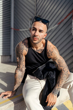 Chico joven musculado y tatuado con ropa urbana posando en la calle frente una pared de chapa gris