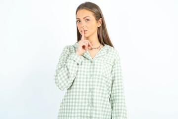 Surprised Young beautiful woman standing over white studio background makes silence gesture, keeps finger over lips and looks mysterious at camera