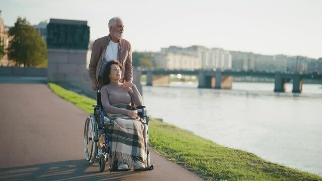Elderly husband walking with wife in wheelchair. Senior family couple spending time in city park. Man caring woman showing love, they have smiling communication. Living with physical disability.