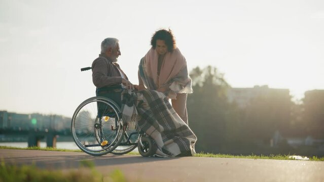 Wife helps husband to sit in wheelchair. Woman putting elderly man legs on wheelchair footplates, walking together in park. Caring female help male with special needs, physical disability concept.