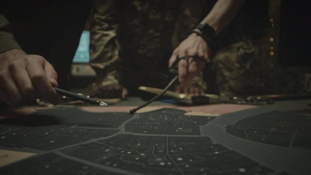 Cropped closeup of unrecognizable military workers with walkie talkies in hands examining points on printed satellite map while co-working at night in office