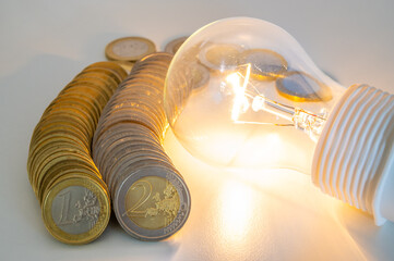 Light bulb lit, with row of coins next to it. Trends in electricity tariffs, energy dependence, energy supplies.