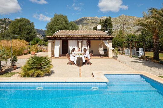 Friends gathered around a rustic table for a delightful outdoor meal by the pool, with a majestic mountain backdrop and lush garden surroundings.