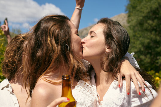 A tender moment of a friendly kiss on lips between two women amidst the joy of a sunny outdoor gathering, with one holding a beer bottle.