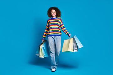 Full length photo of lovely young lady walk hold shopping bags wear trendy rainbow print garment isolated on blue color background