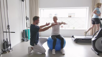 elderly woman sits on a large exercise ball, arms extended, following posture instructions from a physiotherapist