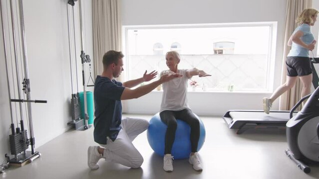 elderly woman sits on a large exercise ball, arms extended, following posture instructions from a physiotherapist