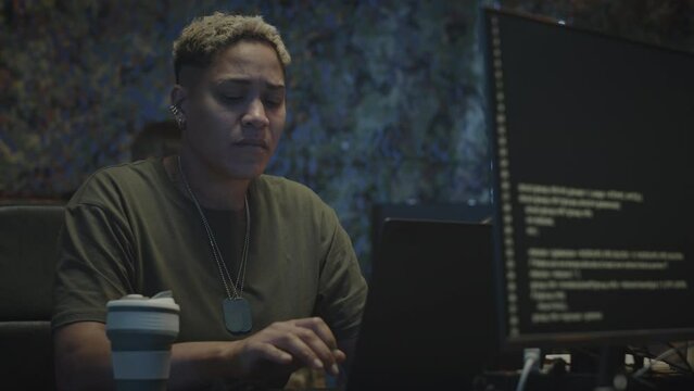 Waist Up Of Biracial Female Military Cyber Security Worker Typing Program Code On Laptop Computer Sitting At Dark Office At Night Time With Colleague