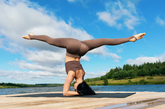 Attractive woman performing Shirsasana exercise with Hanumanasana, inverted asana, headstand with longitudinal splits, training on the shore of a lake on a warm sunny morning