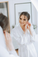 Beautiful, young, perfect model bride in a white lace dress sits in the room, bedroom in the morning, putting an earring on her ear. Wedding photography, portrait, concept.