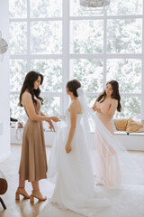 Naklejka premium Wedding morning. Bridesmaids help put on the white wedding dress. A young woman is preparing to meet her groom and having fun with her friends