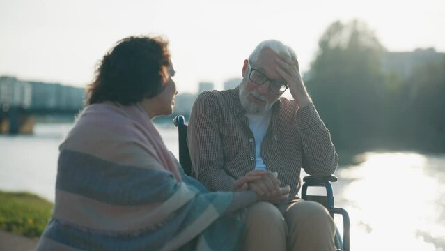Elderly wife walking with husband in wheelchair. Senior family couple spending time in city park. Woman supporting talking with man showing love care. Living with physical disability concept.