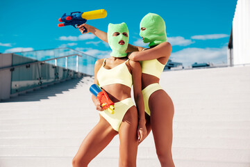 Two beautiful sexy women in green underwear. Models wearing bandit balaclava mask. Hot seductive female in nice lingerie posing in the street at sunny day, blue sky. Crime and violence. Hold water gun