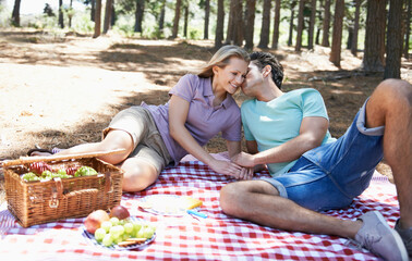 Happy couple, kiss and relax in nature for picnic, love or support in affection, date or outdoor bonding. Woman and man sitting on floor with basket of fruit for embrace, eating or romance in forest