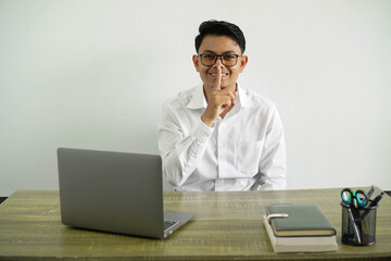 young asian businessman in a workplace showing a sign of silence gesture putting finger in mouth, wearing white shirt with glasses isolated