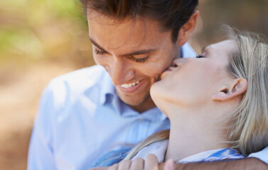 Fototapeta premium Happy couple, kiss and hug in nature for embrace, love or support in outdoor walk or bonding. Young woman and man with smile on cheek for affection, comfort or romance in forest or woods together