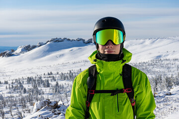 Close-up portrait of a skier or snowboarder in sports equipment, snowy mountains background at ski resort. Bright acid green outfit: warm suit jacket, goggles, black sport helmet, backpack straps - obrazy, fototapety, plakaty