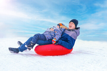Cheerful young man with little boy ride tubing down a hill in the snow in winter. Winter entertainment. Snow fun.Frosty sunny day.Copy space. - obrazy, fototapety, plakaty