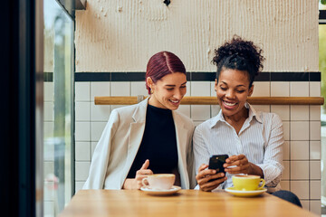 Two happy friends, spending time at the cafe, enjoying their coffee cups, using a mobile phone.