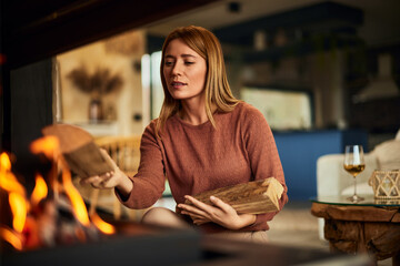 A blonde female holding logs, putting it in the fireplace, at home, during the winter season.