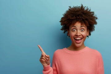 Horizontal shot pleased dark skinned female with Afro haircut, points away with both fore fingers, shows blank space for your promotion, isolated over blue background. People, advertisement concept