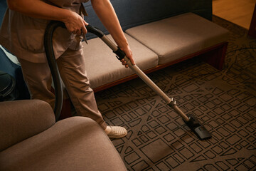 Uniformed chambermaid is vacuuming hotel room with vacuum cleaner