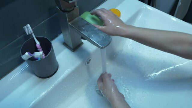 The girl follows proper hand hygiene by thoroughly washing her hands with soap and water in a bathroom sink, a high-angle medium view video of hands, a tap, and a white sink.