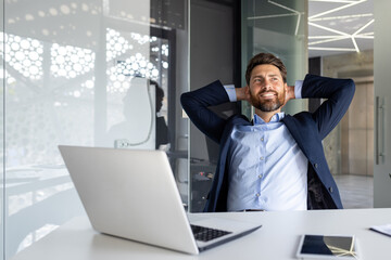 Happy and smiling young businessman man sitting in the office at the desk and resting and satisfied with the work with his hands behind his head