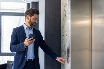 A smiling young man businessman is standing in an office space near the elevator and presses a call button, holding a phone in his hands © Tetiana