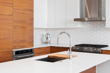 A kitchen faucet detail with modern wood and white cabinets, white marble countertop, stainless...