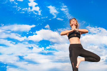 Slender blonde girl doing yoga outside against a blue sky on a sunny day.