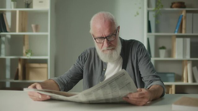 A Serious, Smart Old Man Sits at a Table and Reads a Newspaper.An Elderly Man with a Newspaper Alone at Home, Sees the News and Current Events in the Newspaper.Daily Newspaper Concept