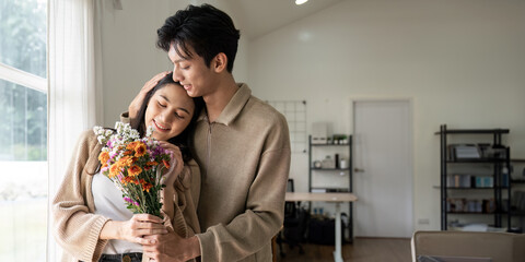 Romantic young asian couple embracing with holding flowers and smiling in living room at home. fall...