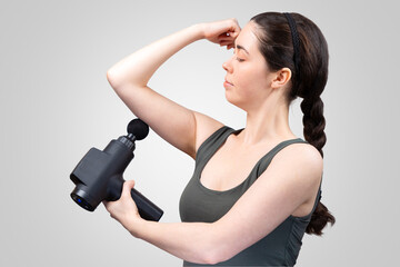 Side view of adult pretty Caucasian woman gives herself massage on arm with percussion massager-gun with ball nozzle. Gray studio background. Therapy for joint pain relief