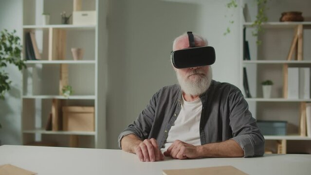 A Modern Old Man With Gray Beard in VR Glasses Examines Everything Around. An Elderly Man in VR Glasses Sitting at a Table at Home.VR to Help the Elderly.