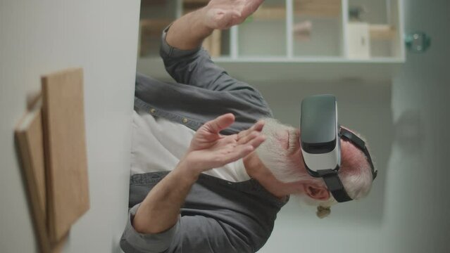 Vertical View.A Modern Old Man in VR Glasses Examines and Touches Everything Around. An Elderly Man in VR Glasses Sitting at a Table at Home.Virtual Reality Games for the Elderly.
