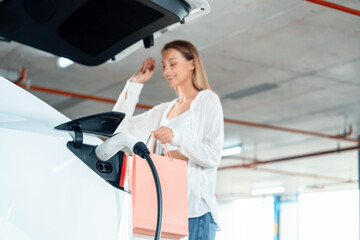 Young woman travel with EV electric car to shopping center parking lot charging in downtown city...