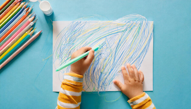 Baby hand holding marker and drawing colorful scratches lines on white paper. Blue table background. Pastel color. Closeup. Toddler development. Learning painting. Point of view shot. Top down view
