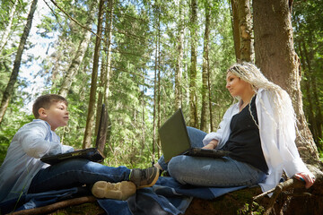 Fototapeta premium Mother and son with a laptops in forest in summer. Fat young smart teenage boy and woman working with modern IT technologies in nature