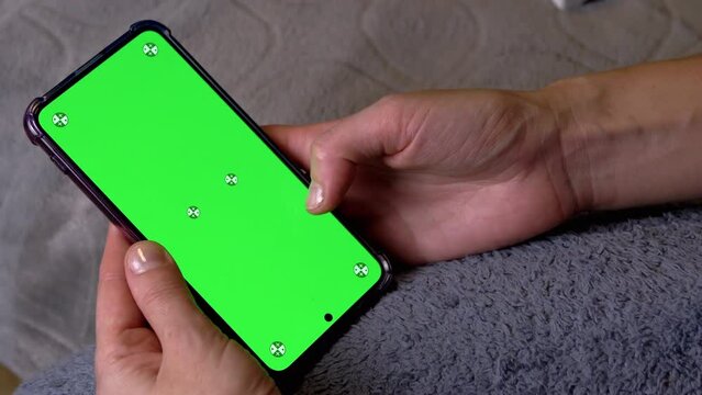 Woman Hand Holding Smartphone with Green Screen in Room at Bedtime. Chroma key layout for copy space. Fingers scrolling through the news feed, video on chromatic screen of the smartphone with fingers.