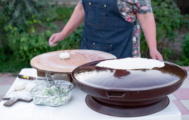 Front view of old female cook wearing apron rolling pita on wooden board, frying on pan outdoor. Concept of fast food.