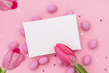 Easter postcard. Invitation mockup or blank with White card for greeting or invitation lies on Easter chocolate pink eggs. Three Red tulips. Shimmering glowing hearts. Happy holidays. Copy space