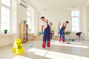 Young team of professional janitors in uniform washing floor with caution wet floor sign at home or...