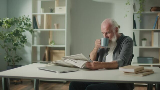 A Serious, Smart Old Man Sits at a Table and Reads a Newspaper.An Elderly Man with a Newspaper, Drinking Coffee Alone at Home, Sees the News and Current Events in the Newspaper.Daily Newspaper Concept