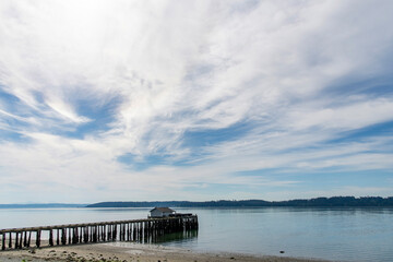 Fototapeta na wymiar Panoramic view over the water of Penn Cove near Coupeville, WA, USA with a wooden fishing dock and building extended in the water against a blue sky with white feather clouds 