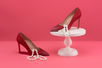 Pair of red high heel shoes with necklace on a stand on pink background.