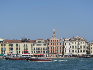 Naklejka premium Brown tourist boats sail along the ancient buildings of Venice, leaving behind traces on the water. Waves on the blue sea