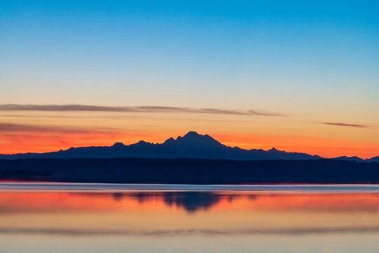 Colorful panoramic view early summer morning sunrise from Coupeville, WA, USA over Skagit Bay towards stratovolcano Mt Baker in Cascades Mountain range in silhouette and reflection in tranquil water