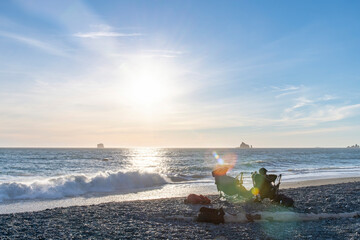 View of two people in chairs on Rialto Beach on coastal stretch of Olympic National Park, WA, USA with lens flare of late afternoon low sun and large number of sea stack rock formations in the ocean © Sonja